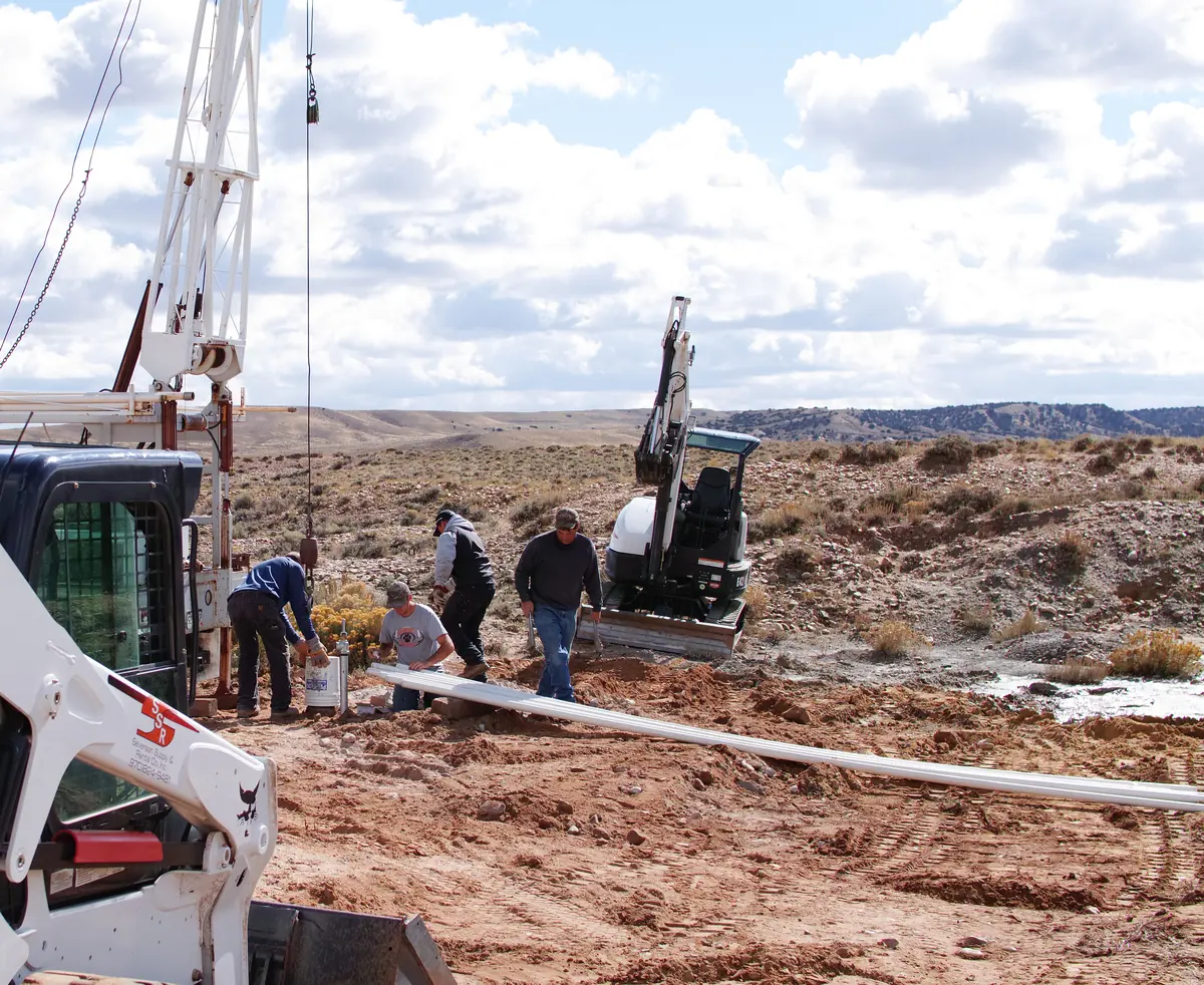 Drilling crew installing the water well at the West Side Placer, Moffat County, Colorado — a drill rig, tracked excavator and skid-steer working shallow sand and gravel in high desert terrain.